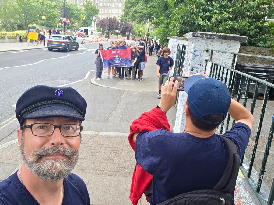 Jeffrey Fabiszewski wearing a navy Breton cap with a Scouting pin at Abbey Road, London, while American Sea Scouts from Ship 611 pose with their flag in the background.