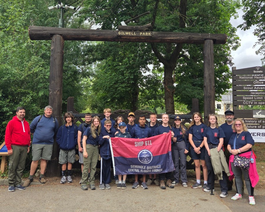 American Sea Scouts from Ship 611 pose beneath the Gilwell Park entrance sign in England, with Jeffrey Fabiszewski wearing a navy mariner's cap at the right